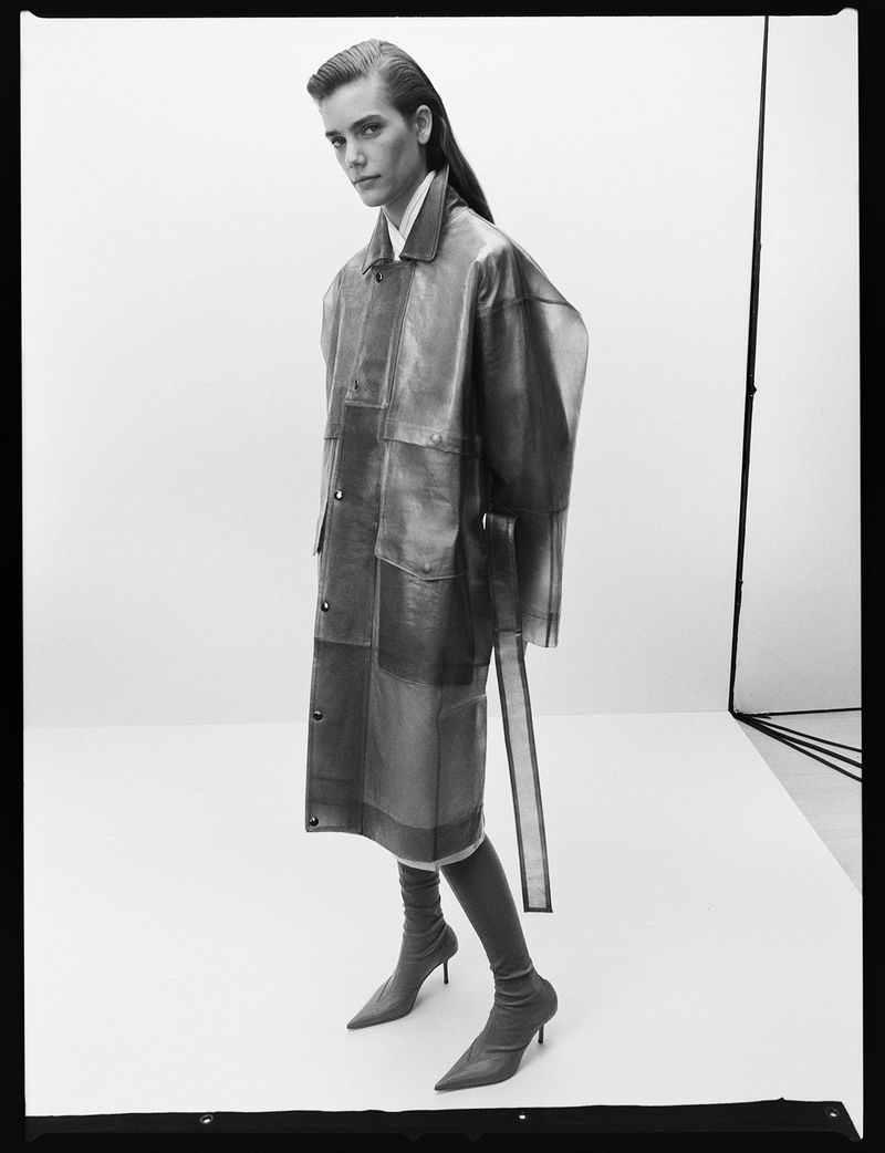 Monochrome portrait of a woman in a leather trench coat and matching boots against a neutral backdrop in a studio setting.