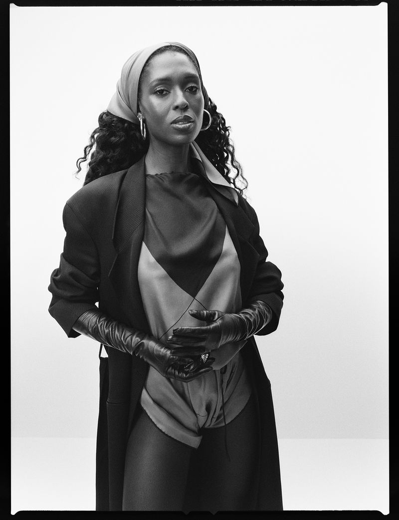 Black and white studio portrait of a Black woman wearing a headscarf, draped jacket, and gloves, with hands clasped.