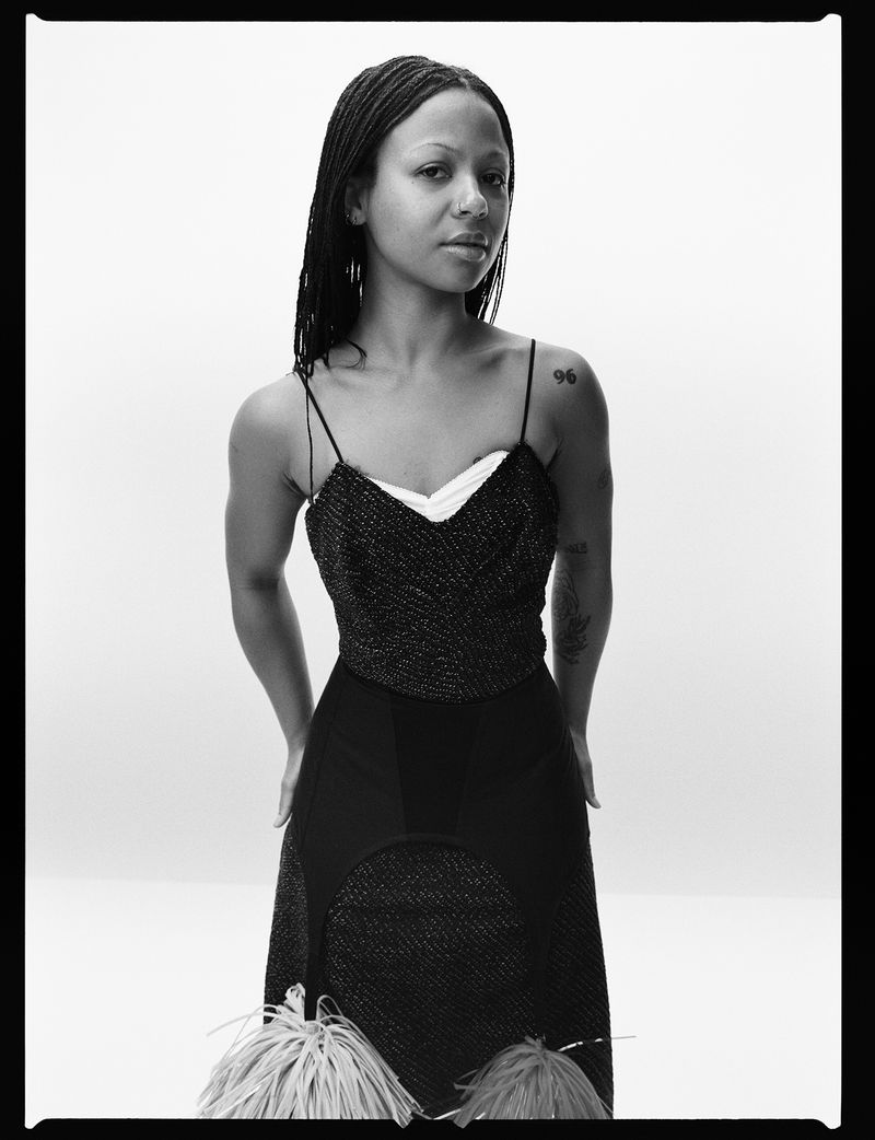 Black and white portrait of a woman with braided hair wearing a spaghetti strap dress with hands on her hips in a photo studio.