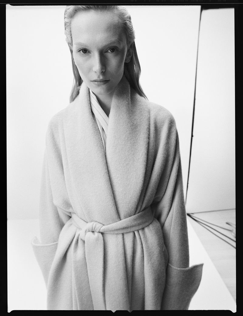 Monochromatic portrait of a woman in a light coat with a tied belt, posing indoors with soft lighting and a neutral background.