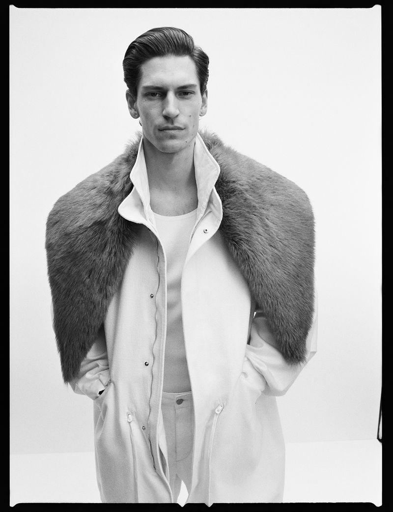 Black and white portrait of a man in a fur stole and light coloured coat posing in a photo studio with soft, neutral lighting.
