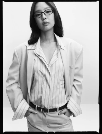 East Asian woman wearing an oversized light blazer, striped shirt, and black belt, looking at the camera against a plain background.