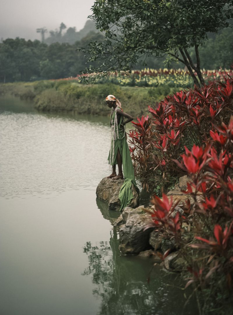 An adult stands by a lake with red plants; trees and foliage are in the background under a cloudy sky creating a peaceful nature scene.
