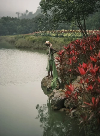 An adult stands by a lake with red plants; trees and foliage are in the background under a cloudy sky creating a peaceful nature scene.