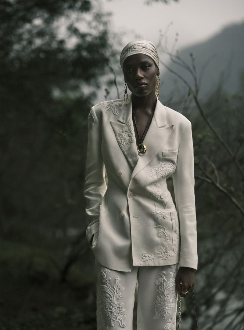 A striking fashion portrait of a Black woman in an embroidered white pantsuit and headscarf, set against a moody, natural background.