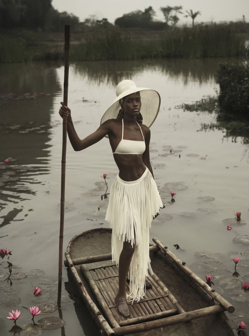 Elegant woman in white bikini and fringe skirt stands in a wooden boat on a lily pond, holding a bamboo pole in natural daylight.