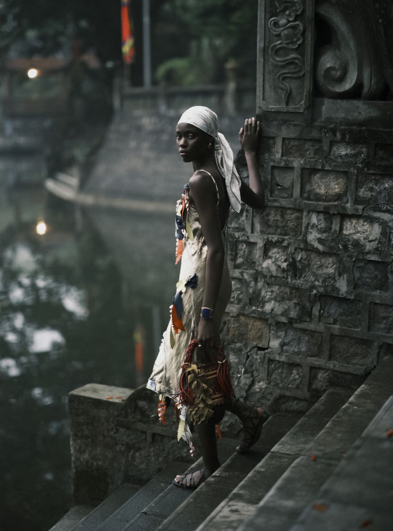 Woman in patterned dress and white headscarf stands on stone steps by a canal, holding a woven basket, outdoor setting, natural light.