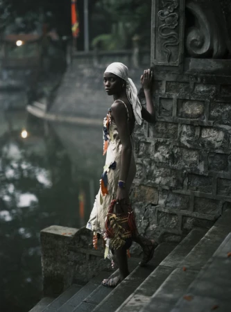 Woman in patterned dress and white headscarf stands on stone steps by a canal, holding a woven basket, outdoor setting, natural light.