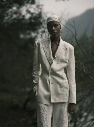 Black woman wearing a white embroidered suit and matching headwrap standing outdoors against a misty mountain background