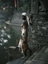 Black woman wearing a headscarf and patterned dress standing on stone stairs next to an ornate wall