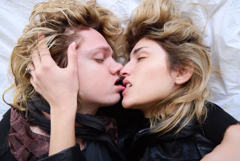 Close-up of a blonde-haired adult man and woman in black jackets intimately touching, with their lips slightly parted against a white background.
