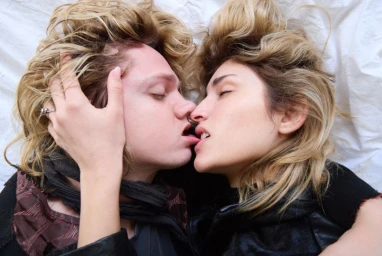 Close-up overhead portrait of a couple kissing passionately while lying on rumpled white fabric background.