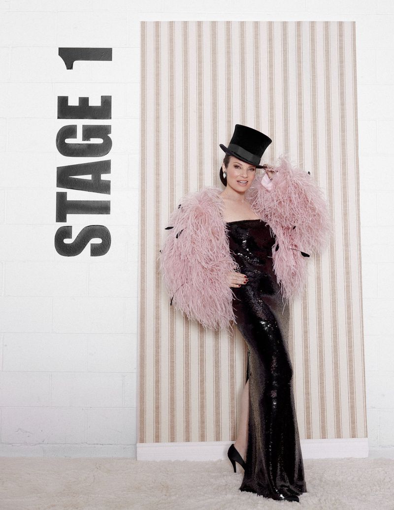 Glamorous woman in black sequin gown and pink feather boa poses confidently against a vertically striped wallpaper and block-lettered wall.