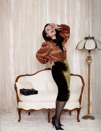 Adult woman strikes a pose in a retro-inspired outfit in a vintage interior with patterned wallpaper, a white sofa, and a floor lamp.