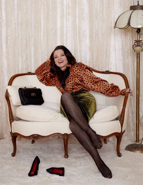 Elegant woman in patterned blouse and velvet skirt poses on a vintage settee, black handbag on the cushion, red shoes on floor.
