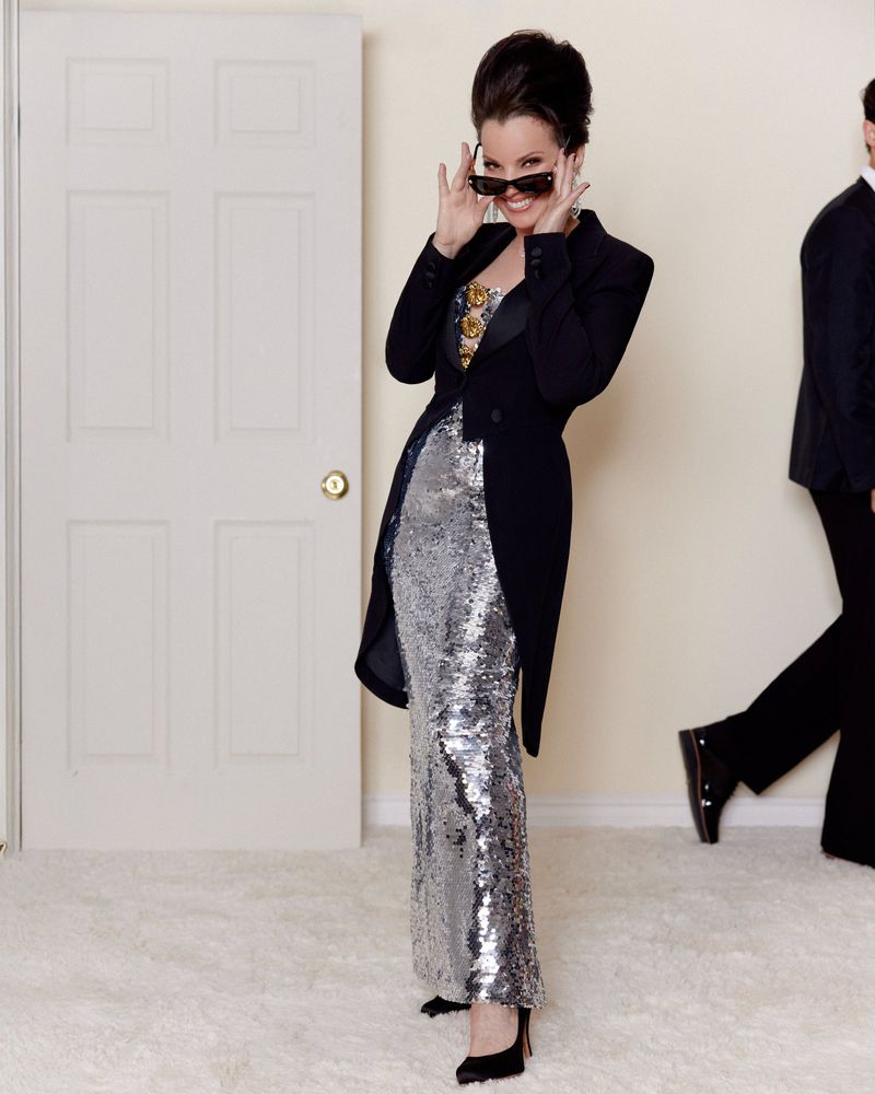 Stylish woman in a shimmering silver dress and black jacket poses indoors, peering over sunglasses with a man in the background.