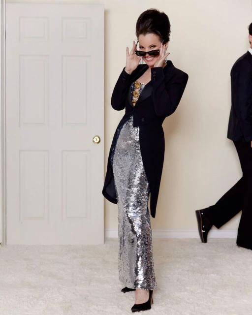 Stylish woman in a shimmering silver dress and black jacket poses indoors, peering over sunglasses with a man in the background.