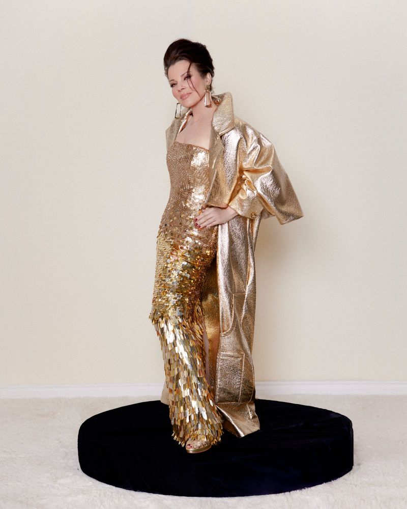 Woman in a shimmering gold gown and long jacket poses on a round, black platform in a studio with an off-white background and fluffy white carpet.