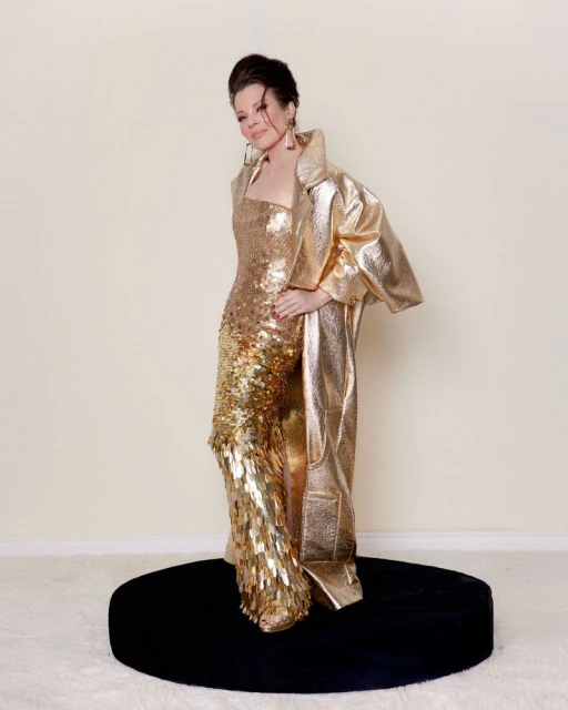 Woman in a shimmering gold gown and long jacket poses on a round, black platform in a studio with an off-white background and fluffy white carpet.