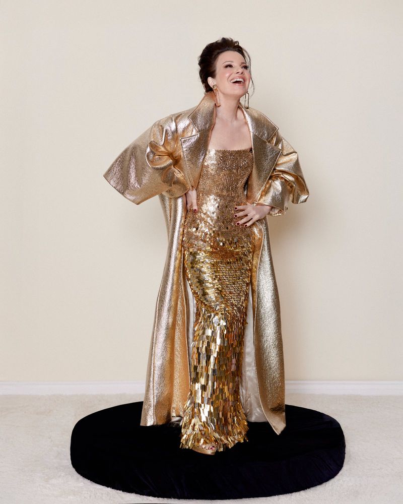 A full shot of a smiling woman in a gold gown and coat stands on a black circular platform against a cream-colored backdrop. 