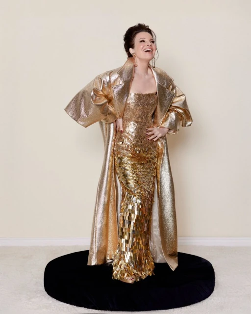A full shot of a smiling woman in a gold gown and coat stands on a black circular platform against a cream-colored backdrop. 