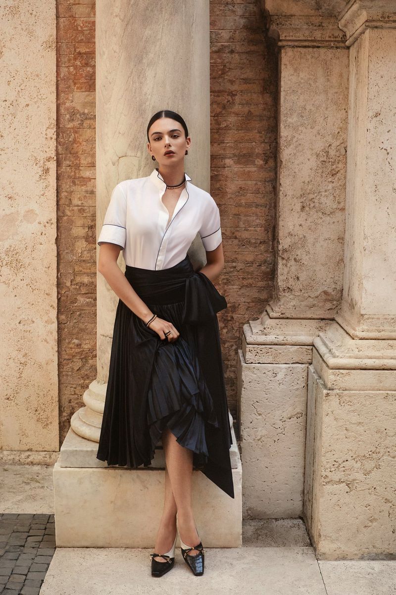 Elegant woman in a white blouse and black skirt posing next to stone pillars in a sunlit outdoor setting for a fashion editorial.