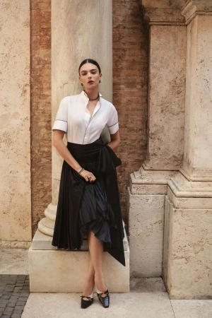 Deval Cassel leaning against a classical stone column in a white short-sleeve shirt and black pleated skirt for iO Donna.