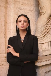 Model Deva Cassel wearing a black blazer and necklace posing against a classical stone column for iO Donna.
