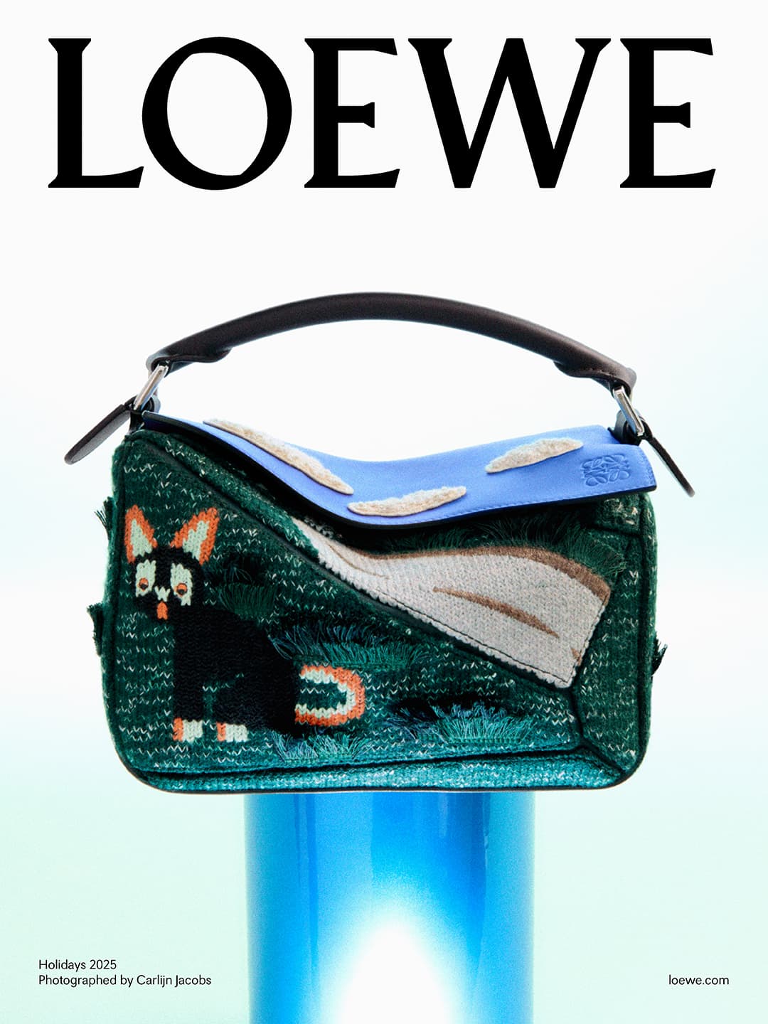 Close-up of a Loewe Puzzle bag with a whimsical cat design, resting on a shiny blue pedestal against a soft, gradient backdrop. 