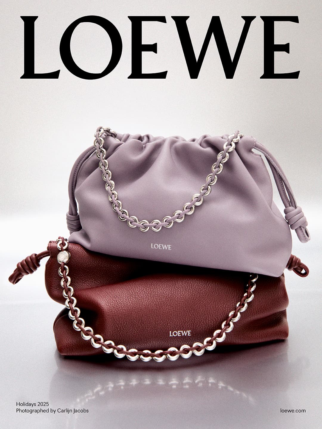 Advertisement for LOEWE showcasing two leather handbags stacked. Each bag is draped with a silver-tone chain. The handbags are lavender and burgundy.