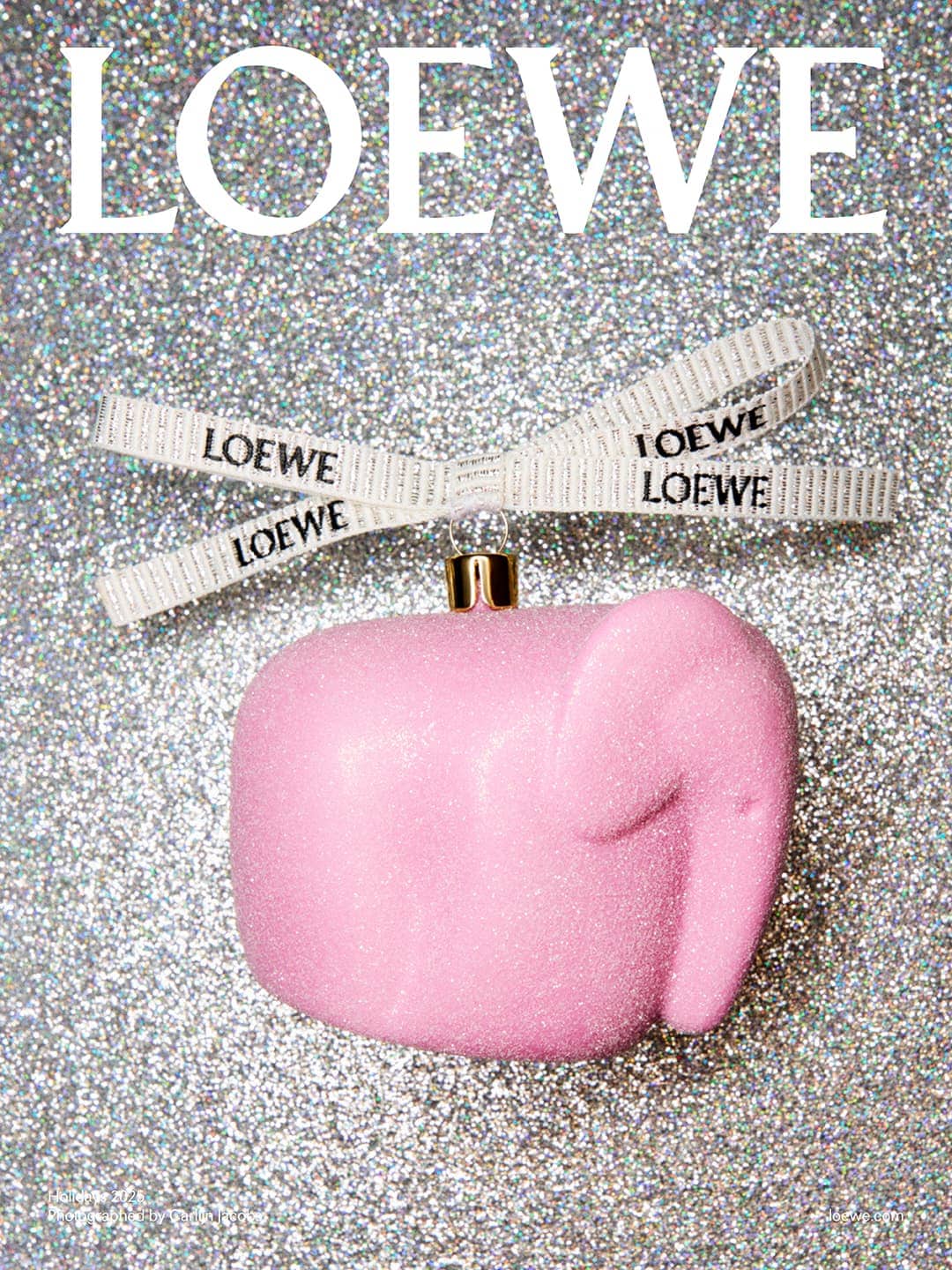 Pink elephant ornament hanging from a white ribbon with the LOEWE logo, set against a silver glittery background. Fashion advertisement.
