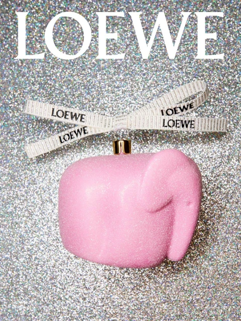Pink elephant ornament hanging from a white ribbon with the LOEWE logo, set against a silver glittery background. Fashion advertisement.