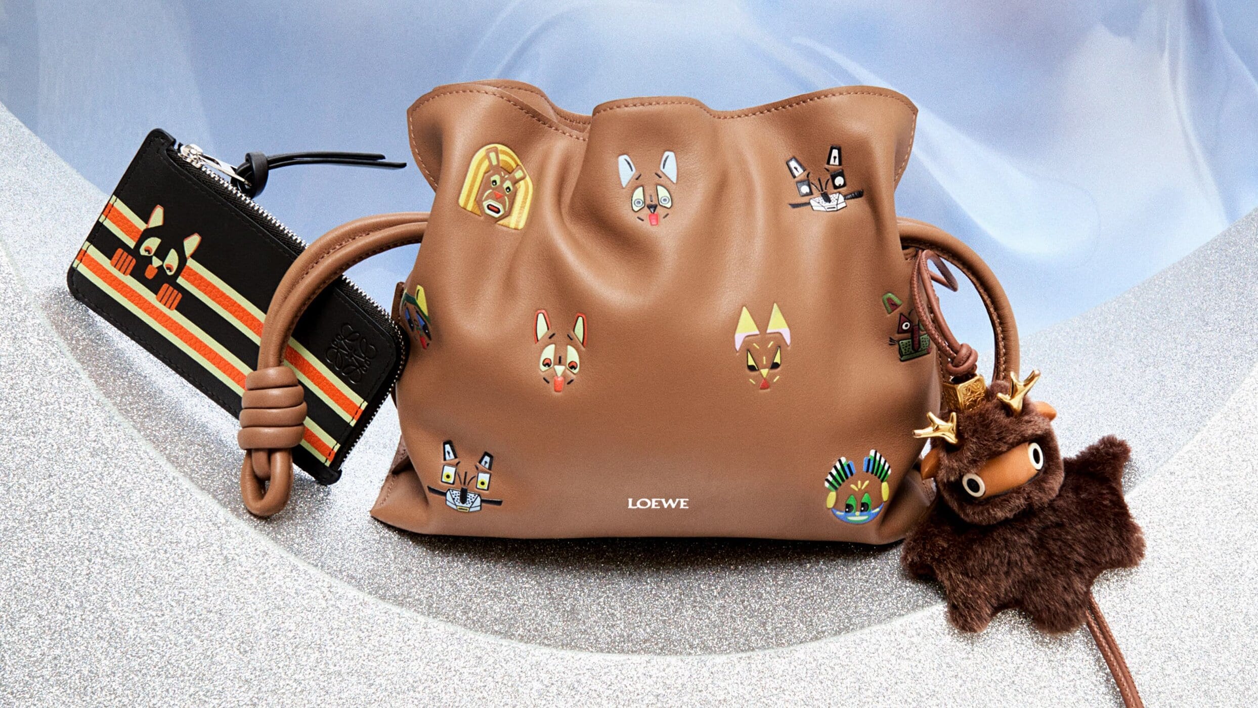 Brown leather pouch bag with cartoon dog face illustrations and a plush dog charm, presented with a black wallet. Fashion still life.