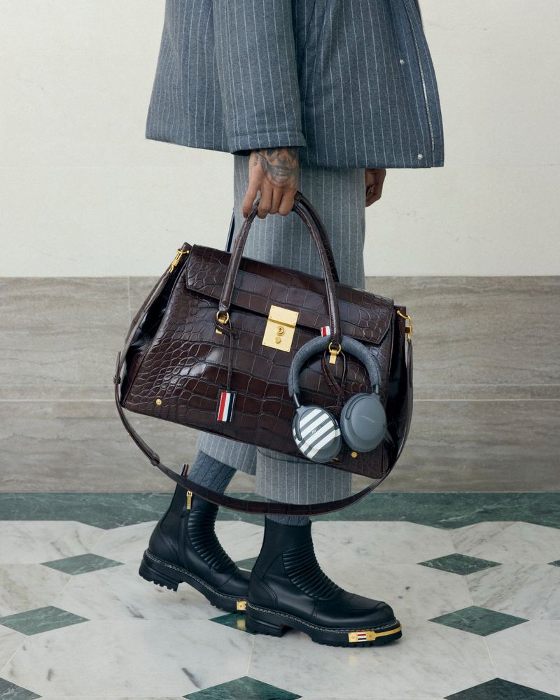 Person carries a brown leather bag and gray headphones, wearing a gray suit, gray socks, and black leather boots with gold details.