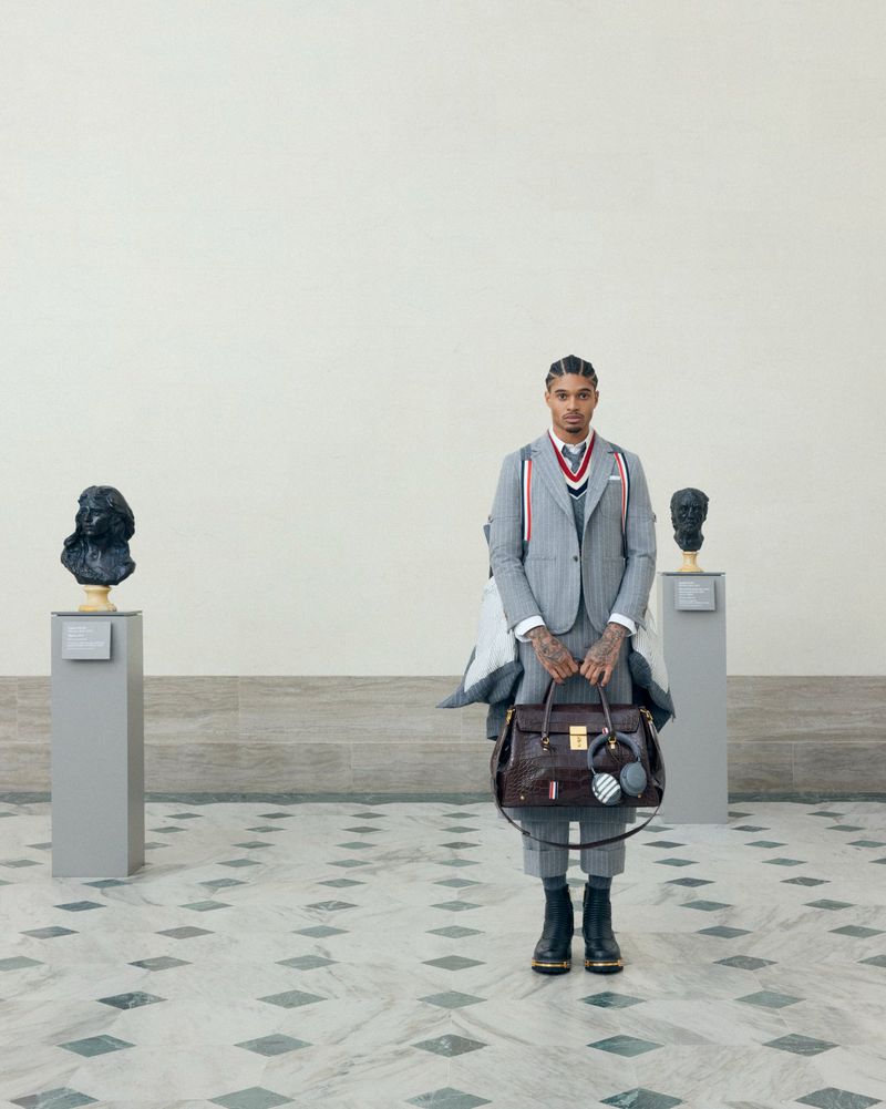 Man in a grey suit stands in an art gallery with busts on pedestals, holding a brown leather satchel and wearing black boots.