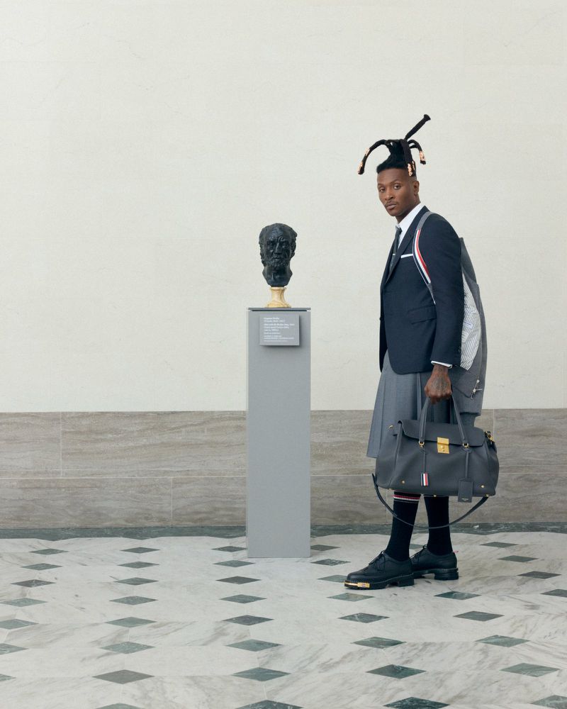 An adult male wearing a black jacket, pleated grey skirt, and black shoes stands next to a sculpture head in an indoor setting.