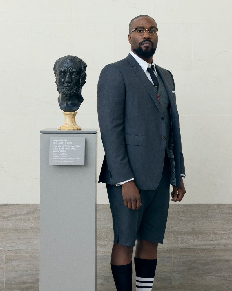 Adult man poses in a grey suit with shorts next to a bust on a grey pedestal, inside a building with white walls and stone floors.