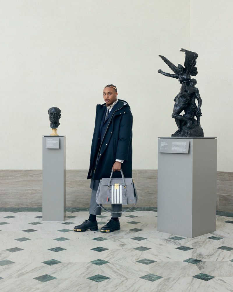 Man in overcoat and grey suit trousers carrying a striped handbag, posing between two sculptures in an art gallery with marble floor.