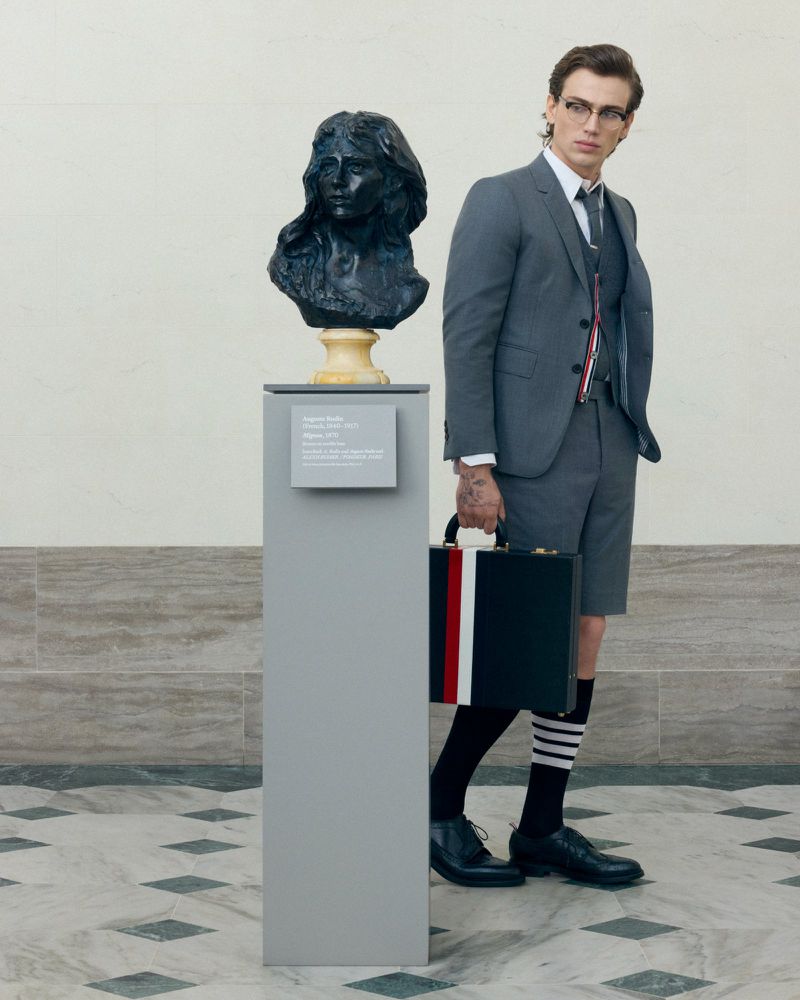 An adult male in a grey suit and striped socks stands next to a bust sculpture on a grey pedestal inside a marble-floored building.
