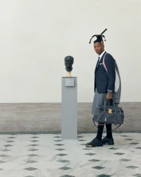 Black man wearing a tailored suit and skirt holding a handbag in a gallery beside a bust on a pedestal