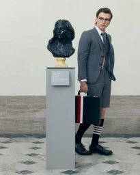 Man in a grey suit and shorts standing beside a museum pedestal with a dark bust and holding a striped briefcase