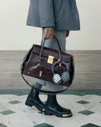 Person wearing grey pinstripe suit and black boots holding a large dark brown crocodile embossed leather handbag with headphones.