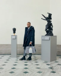 Man holding a grey striped travel bag standing in a museum gallery between two classical statues on pedestals