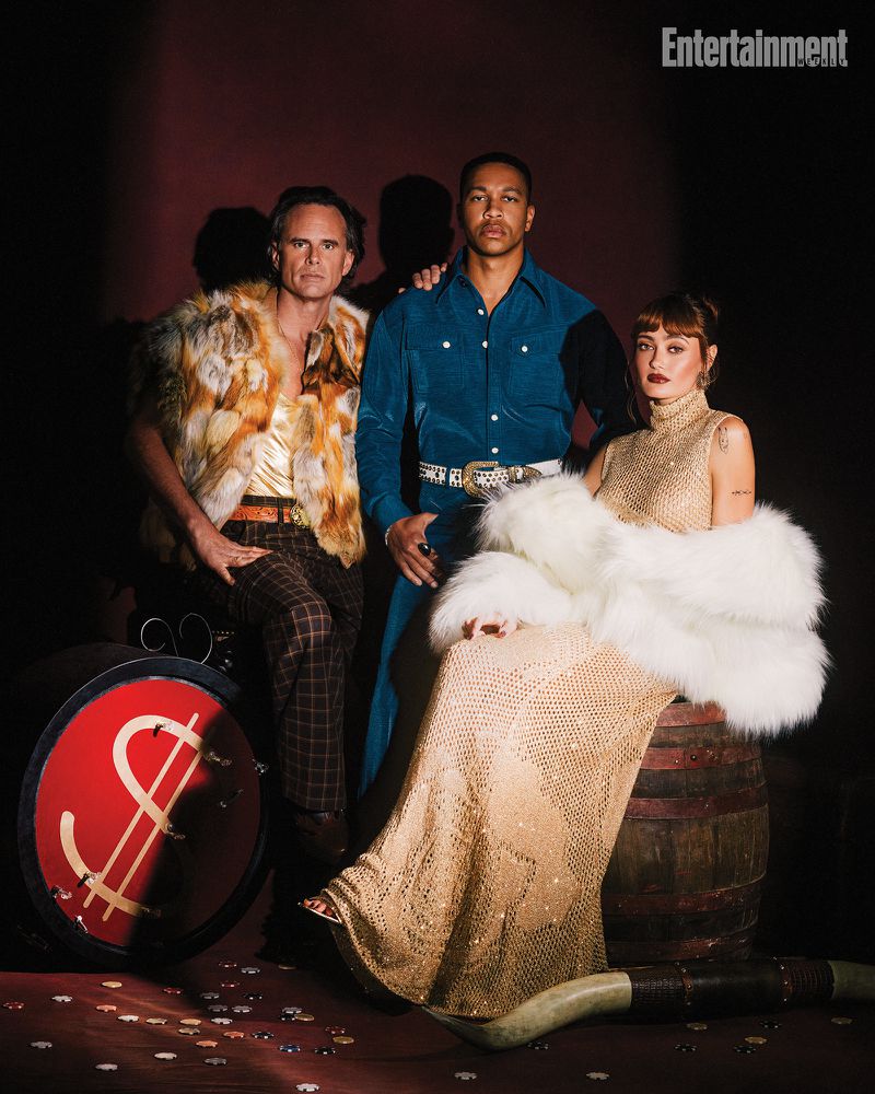 Three figures pose in a vintage-inspired setting, one in a fur vest, another in a blue western shirt, and one in a sequined gown with a fur stole.