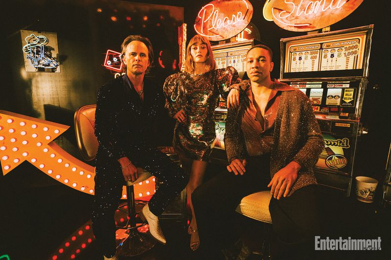 Three people pose indoors in a retro scene featuring a slot machine, illuminated signs, and vintage decor in a warmly lit setting.
