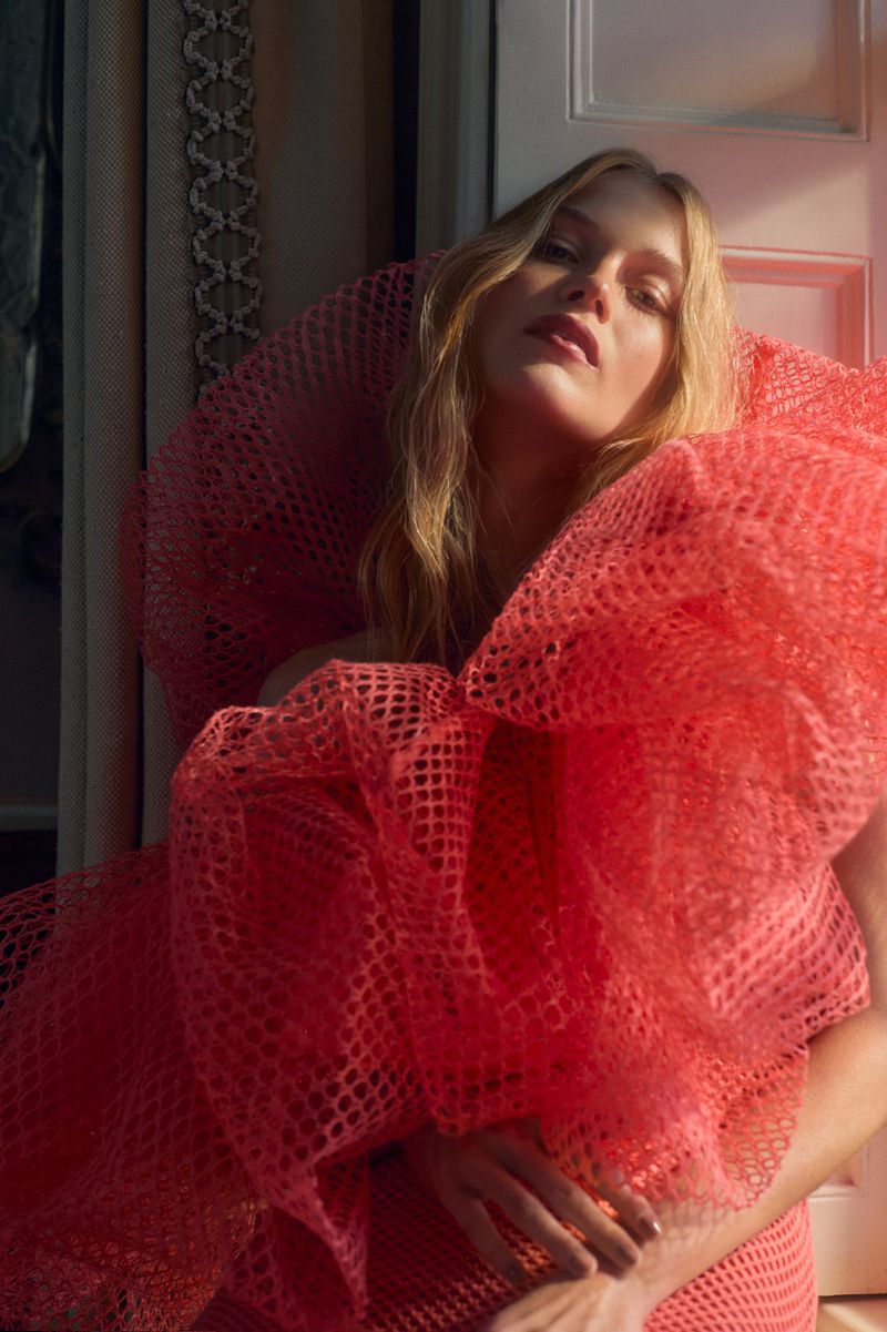 Blonde woman in a large, coral, mesh dress poses indoors with soft, diffused lighting. The model's gaze is upwards, creating an ethereal vibe.