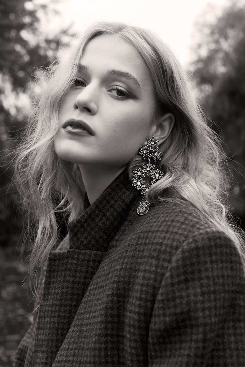 Monochrome portrait of a blonde woman wearing a wool coat and large, decorative earrings against an outdoor, natural background.