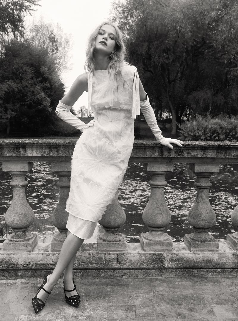 Elegant adult woman in white dress, long gloves, and heels posing on stone balcony with lush background in black and white photography.