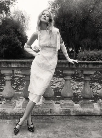 Model in white dress and long gloves leaning against a stone balustrade in a park