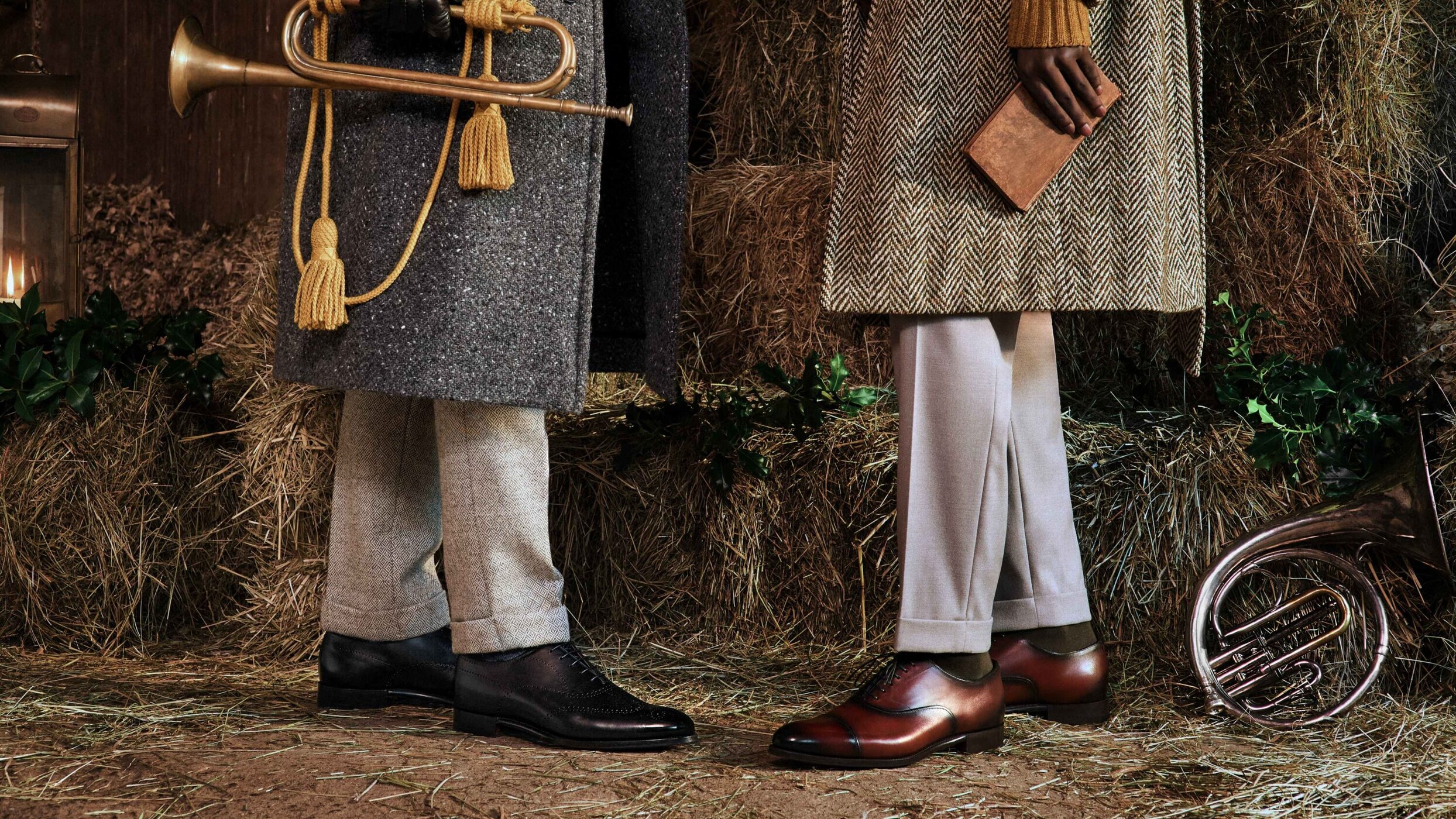 Two people stand in front of hay bales, wearing coats, trousers, and dress shoes. One holds a book, the other a trumpet.